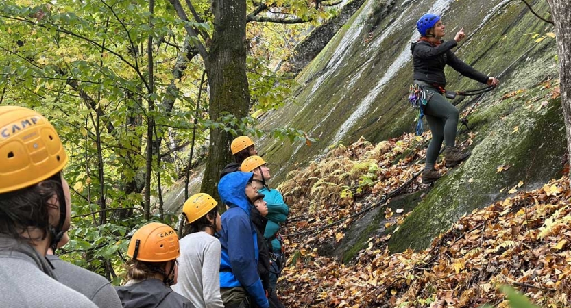 A group of students wearing helmets watch an instructor teach during a rock climbing session 